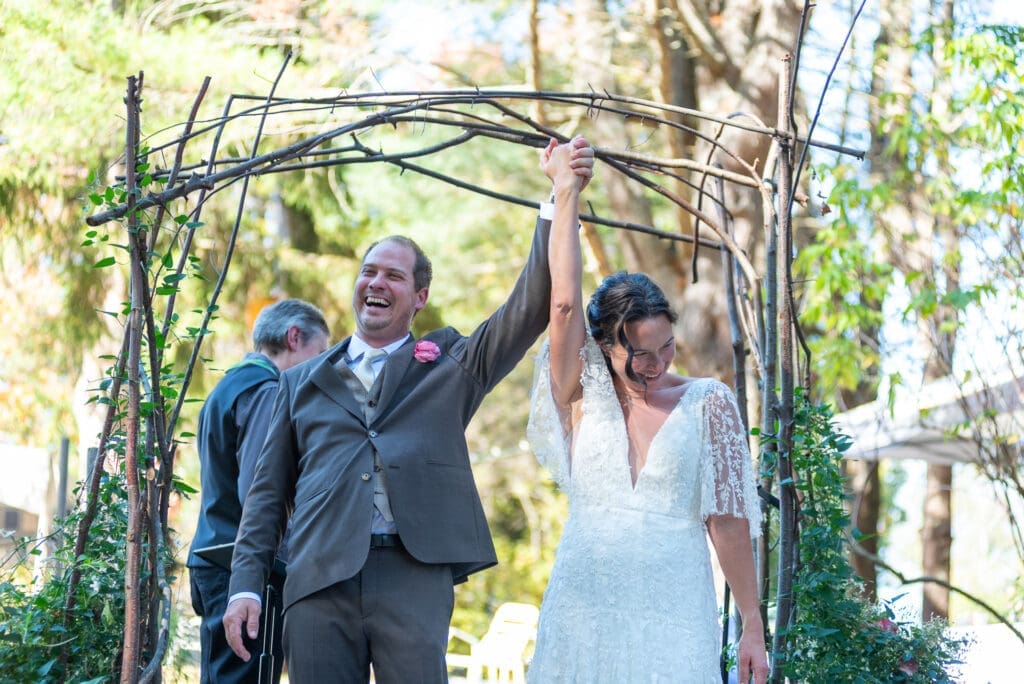 A bride and groom smile and raise their arms as they look out at their guests following their backyard wedding ceremony in Freeport, Maine.