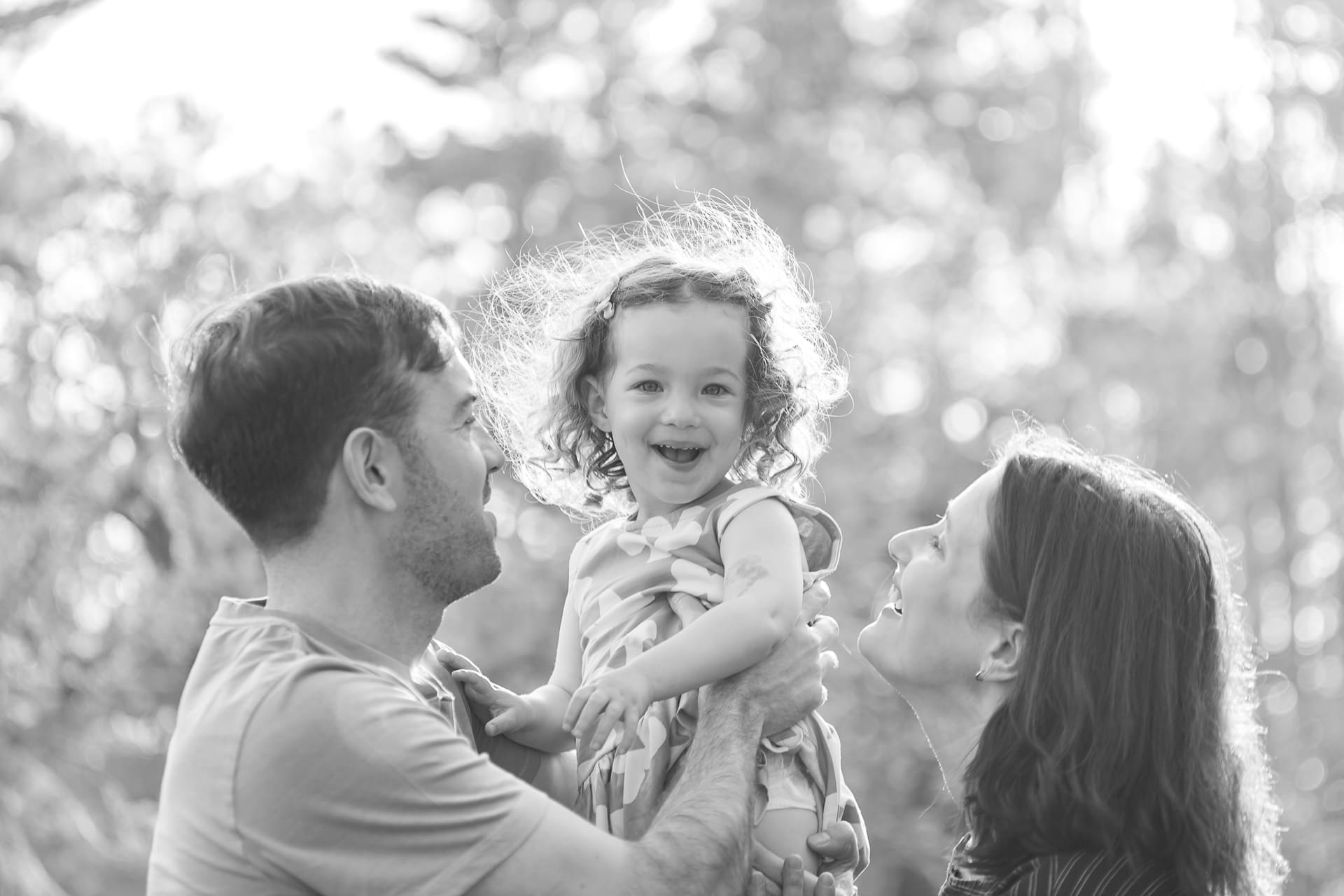 Black and white photograph of a toddler being held in the air by her father while her mother looks on.